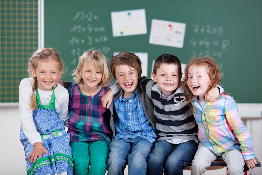 Das Foto zeigt eine Gruppe von lachenden Schulkindern die gemeinsam in einem Klassenraum vor einer Tafel sitzen und dient als Beitragsbild für JUNIOR SERVE Catering Equipment - Das professionelle All-in-One Serviersystem für Kitas und Schulen.