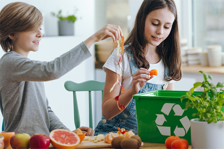 Das Bild zeigt zwei Kinder, die Obst Schälen und die Schalen in einen Recycling-Eimer werfen und ist das Beitragsbild zum Beitrag 'Foodwaste und Müll im Schul- und Kita-Catering vermeiden' auf der Website von JUNIOR-SERVE.
