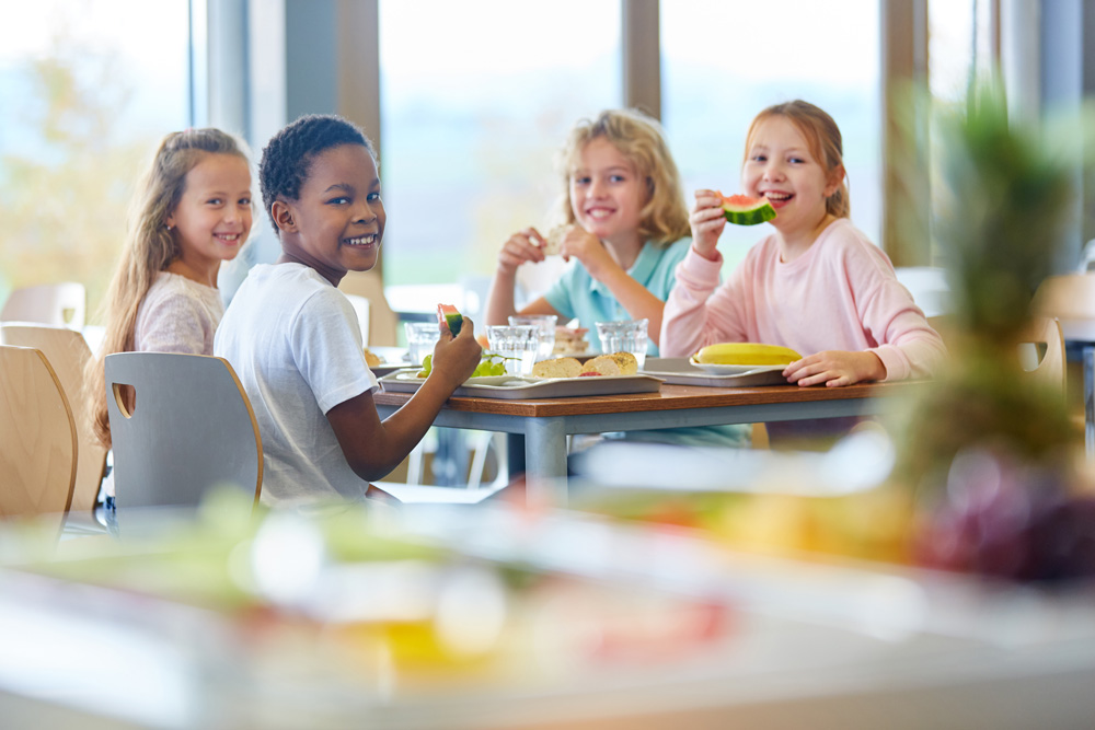 Das Foto zeigt glückliche und lachende Kindern die gemeinsam an einem Tisch essen und soll verdeutlichen, wie einfach die Ganztagsversorgung an Kitas und Schulen mit dem Cook and Chill Verfahren sowie Catering Equipment von JUNIOR-SERVE ist.