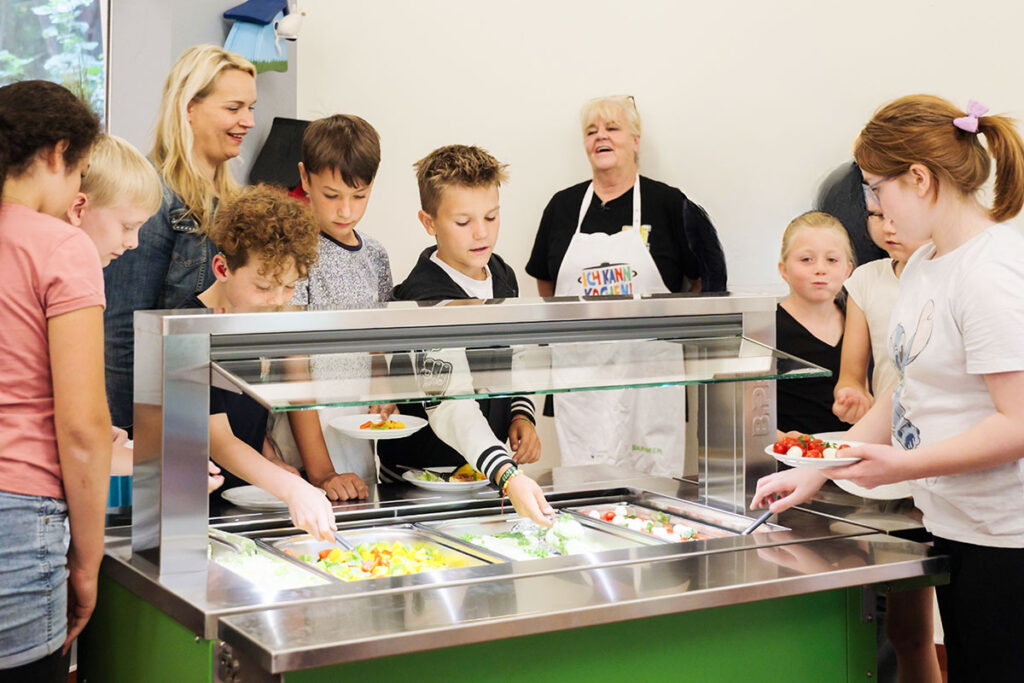 Schüler*innen der Schule stehen in der Mensa oder im Speiseraum, die Ausgabe läuft – sichtbar ist das kindgerechte Ambiente und der Fokus auf Gemeinschaft beim Mittagessen.