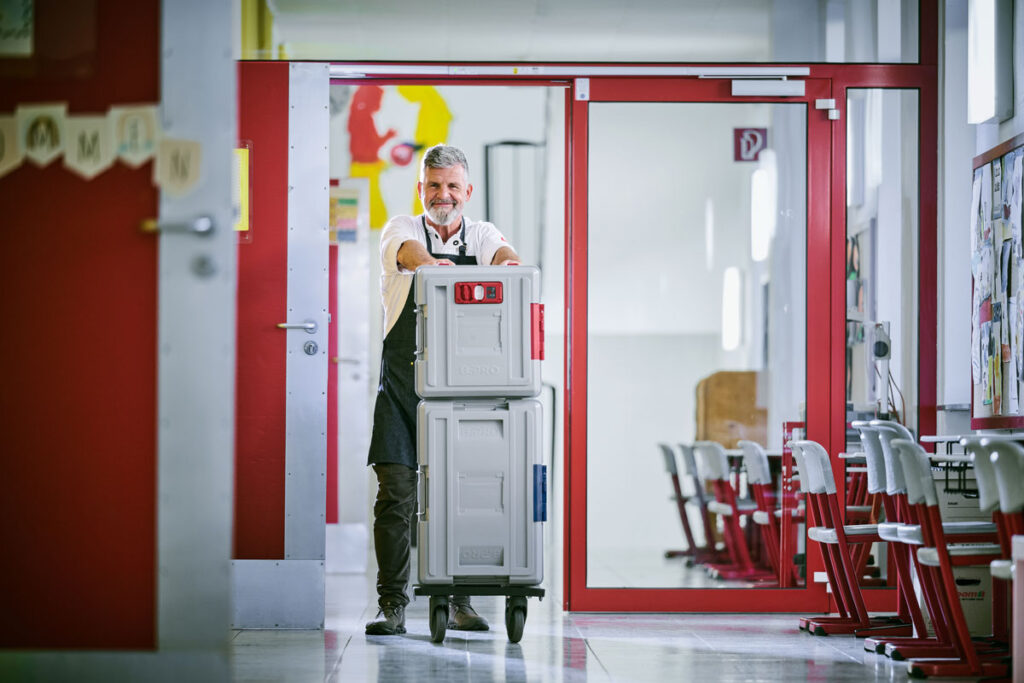 Mitarbeiter schiebt einen grauen B.PROTHERM-K-Kunststoffbehälter durch einen Flur in einer Schule.